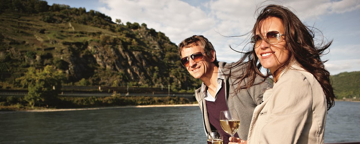 Zwei Personen stehen auf dem Sonnendeck eines Flusskreuzfahrtschiffs und genießen die Aussicht auf den Rhein bei einem Glas Weißwein, umgeben von bewachsenen Hügeln und blauem Himmel.