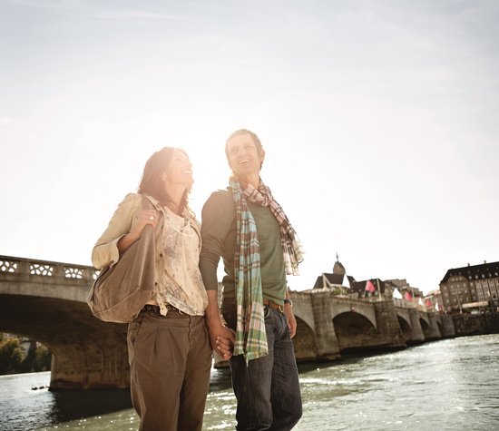 Ein Paar spaziert in Basel am Ufer des Rheins bei Sonnenschein entlang. Im Hintergrund ist eine steinere Brücke, die über den Fluss führt.