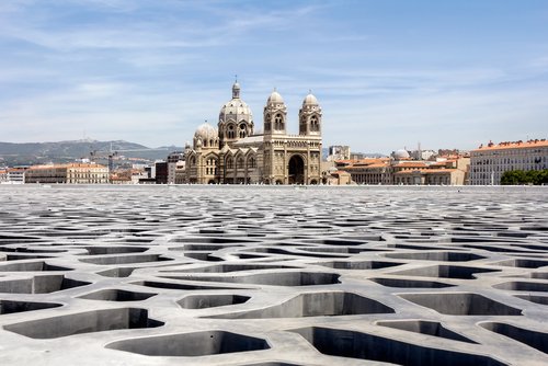 Die Kathedrale La Major in Marseille hebt sich mit ihren markanten Kuppeln vom hellen Himmel ab, davor breitet sich das moderne Dach des Museums MuCEM aus.