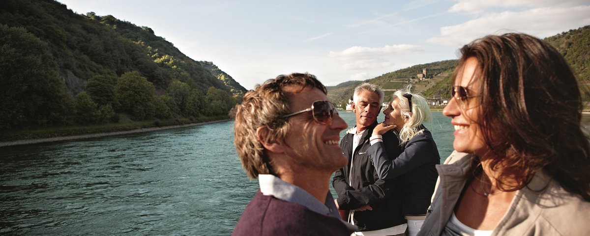 Zwei Paare stehen auf dem Sonnendeck eines Flusskreuzfahrtschiffs, genießen die Aussicht auf den Rhein und die umliegende Hügellandschaft bei sonnigem Wetter.