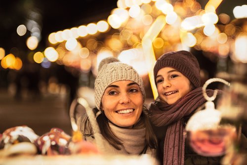 Frau und Kind mit Mütze und Schal lächeln auf einem Weihnachtsmarkt bei Nacht mit Lichterketten im Hintergrund.