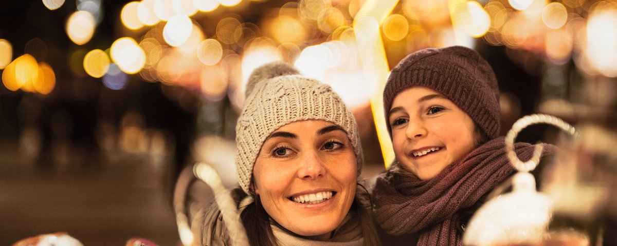 Frau und Kind mit Mütze und Schal lächeln auf einem Weihnachtsmarkt bei Nacht mit Lichterketten im Hintergrund.