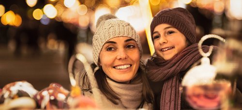 Frau und Kind mit Mütze und Schal lächeln auf einem Weihnachtsmarkt bei Nacht mit Lichterketten im Hintergrund.