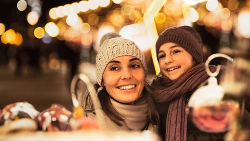 Frau und Kind mit Mütze und Schal lächeln auf einem Weihnachtsmarkt bei Nacht mit Lichterketten im Hintergrund.