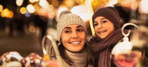 Frau und Kind mit Mütze und Schal lächeln auf einem Weihnachtsmarkt bei Nacht mit Lichterketten im Hintergrund.