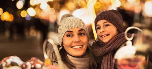 Frau und Kind mit Mütze und Schal lächeln auf einem Weihnachtsmarkt bei Nacht mit Lichterketten im Hintergrund.
