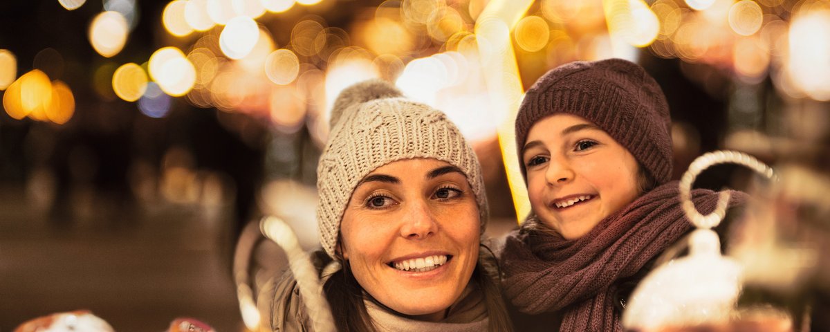 Frau und Kind mit Mütze und Schal lächeln auf einem Weihnachtsmarkt bei Nacht mit Lichterketten im Hintergrund.