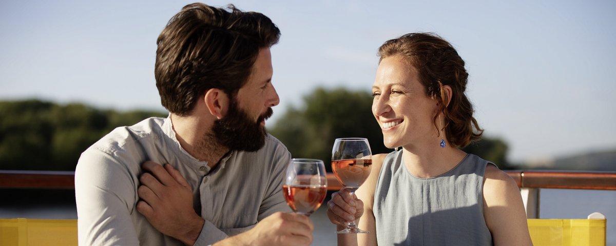 Paar sitzt draußen und stößt mit Gläsern Roséwein an, auf dem Tisch Brot und Käse mit Trauben.
