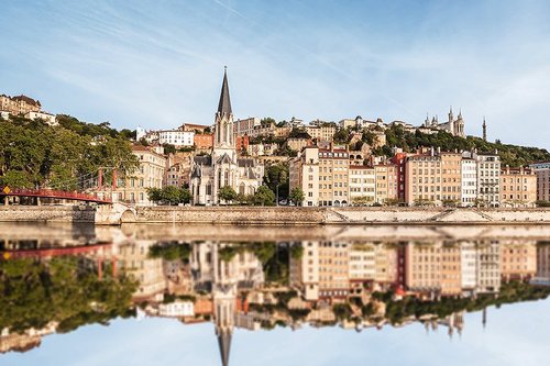 Eine Kirche mit spitzem Turm, eine Brücke über die Rhône und die umliegenden Altbauten spiegeln sich leicht im ruhigen Flusswasser von Lyon.