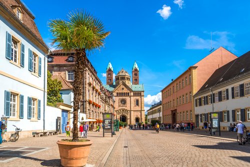 Der Dom zu Speyer unter klarem blauen Himmel, davor eine Straße mit Fußgängern, historischen Gebäuden und Haltestellen bei Sonnenschein.