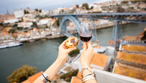 Zwei Hände halten ein Glas Portwein und ein Pastel de Nata vor der Ponte Dom Luís I Brücke in Porto, Portugal