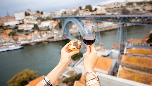Zwei Hände halten ein Glas Portwein und ein Pastel de Nata vor der Ponte Dom Luís I Brücke in Porto, Portugal