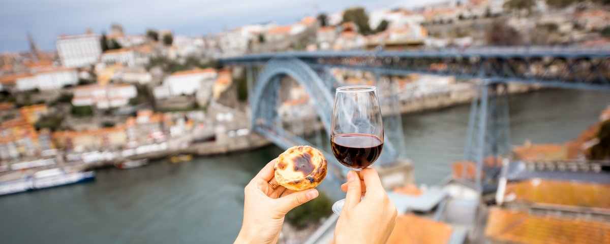 Zwei Hände halten ein Glas Portwein und ein Pastel de Nata vor der Ponte Dom Luís I Brücke in Porto, Portugal