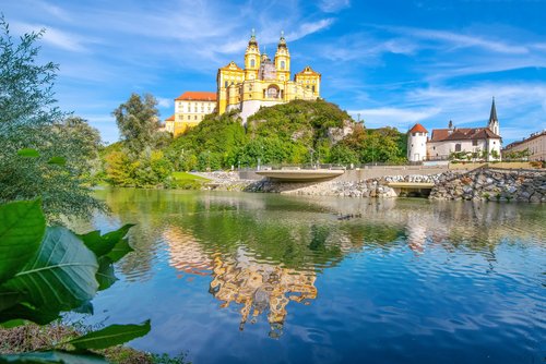 Blick auf das barocke Stift Melk in Österreich, umgeben von üppigem Grün und der Donau im Vordergrund, unter blauem Himmel.