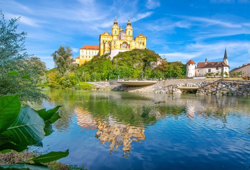 Blick auf das barocke Stift Melk in Österreich, umgeben von üppigem Grün und der Donau im Vordergrund, unter blauem Himmel.