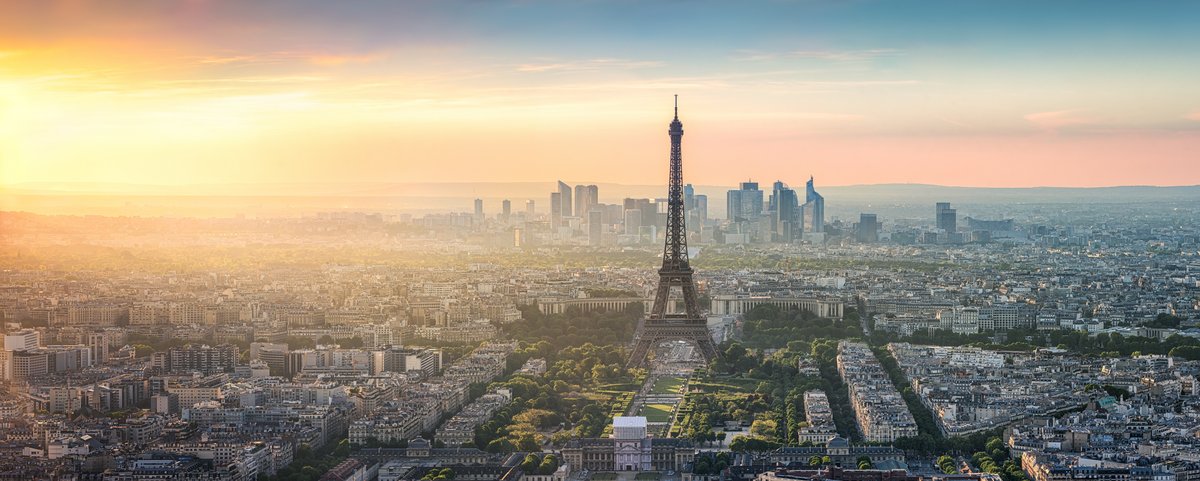 Blick auf den Eiffelturm in Paris bei Sonnenuntergang, umgeben von Bäumen und Gebäuden, mit einem klaren Himmel im Hintergrund.