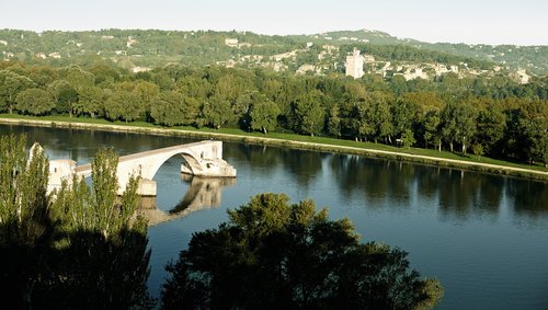 Die mittelalterliche Brücke Pont Saint-Bénézet in Avignon umgeben von einer grünen Flusslandschaft und blauem Himmel.