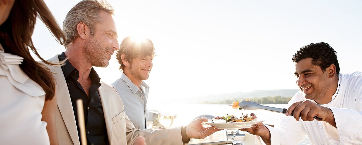 Drei Personen stehen beim Barbecue auf dem Sonnendeck eines Flusskreuzfahrtschiffes und der Koch füllt ihnen Essen vom Grill auf, im Hintergrund der Fluss und Bäume.