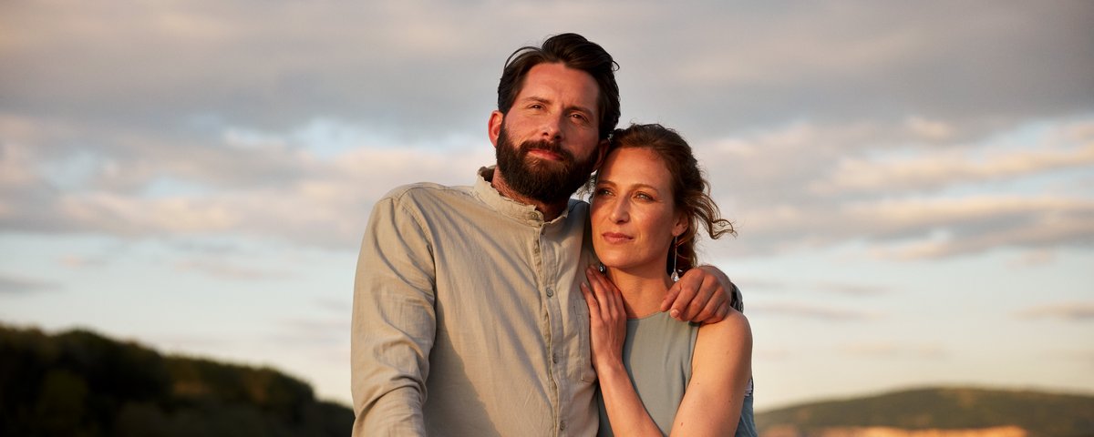 Paar umarmt sich auf einem Boot bei Abendlicht mit Fluss und bewaldeten Hügeln im Hintergrund.