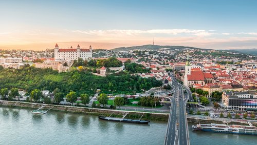 Panorama von Bratislava mit der Donau und einer Brücke im Vordergrund. Im Hintergrund Burg Bratislava auf einem Hügel, umgeben von der Stadt bei schönem Wetter.