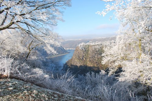 Winterliche Landschaft mit der Loreley am Rhein, schneebedeckte Hügel und der Fluss im Vordergrund unter einem klaren blauen Himmel.