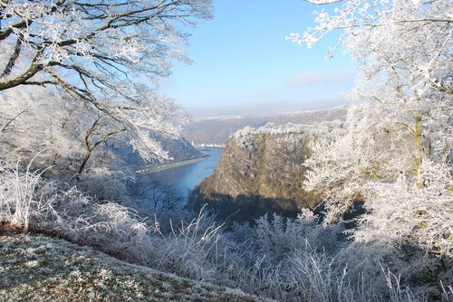 Winterliche Landschaft mit der Loreley am Rhein, schneebedeckte Hügel und der Fluss im Vordergrund unter einem klaren blauen Himmel.