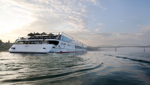 Heckansicht des Flusskreuzfahrtschiffs A-ROSA MIA auf der Donau bei Sonnenaufgang. Im Vordergrund ist der Fluss mit leichten Wellen. Im Hintergrund liegt eine Brücke.