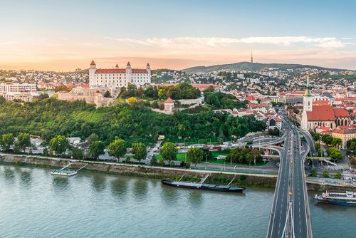 Panorama von Bratislava mit der Donau und einer Brücke im Vordergrund. Im Hintergrund Burg Bratislava auf einem Hügel, umgeben von der Stadt bei schönem Wetter.