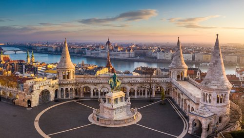 Blick auf die historische Fischerbastei in Budapest mit einer Statue eines Ritters im Vordergrund. Im Hintergrund ein Blick auf das Parlamentsgebäude von Budapest und über die Stadt bei Sonnenuntergang.