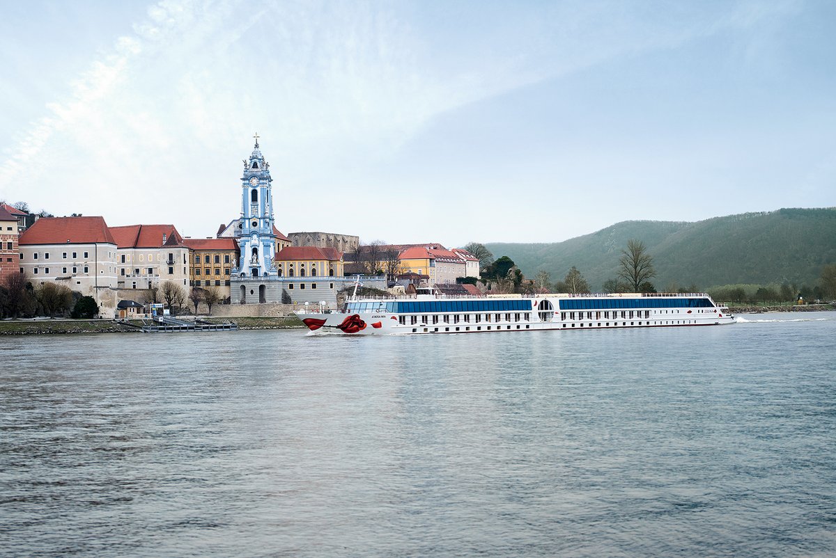 Das Flusskreuzfahrtschiff A-ROSA MIA fährt an Stift Dürnstein auf der Donau vorbei. Im Vordergrund der Fluss, im Hintergrund bewachsene Hügellandschaft unter klarem Himmel.