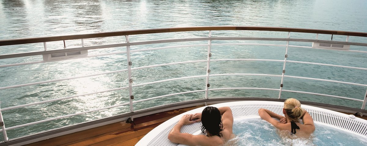 Zwei Personen entspannen in einem Whirlpool auf dem Deck eines Flusskreuzfahrtschiffs mit Ausblick auf den Rhein bei Sonnenschein.