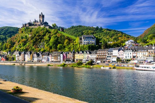 Blick auf die malerische Stadt Cochem mit der Reichsburg auf einem Hügel, umgeben von grünen Weinbergen und dem Fluss Mosel im Vordergrund.