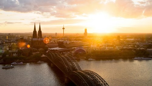 Panoramablick auf die Stadt Köln mit dem Kölner Dom bei Sonnenuntergang. Im Vordergrund die Hohenzollernbrücke, die über den Rhein führt.
