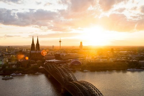 Panoramablick auf die Stadt Köln mit dem Kölner Dom bei Sonnenuntergang. Im Vordergrund die Hohenzollernbrücke, die über den Rhein führt.