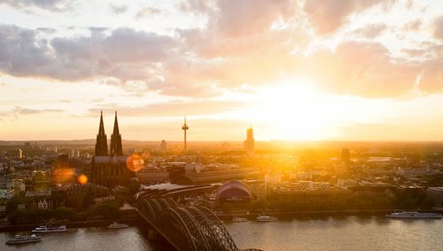 Panoramablick auf die Stadt Köln mit dem Kölner Dom bei Sonnenuntergang. Im Vordergrund die Hohenzollernbrücke, die über den Rhein führt.
