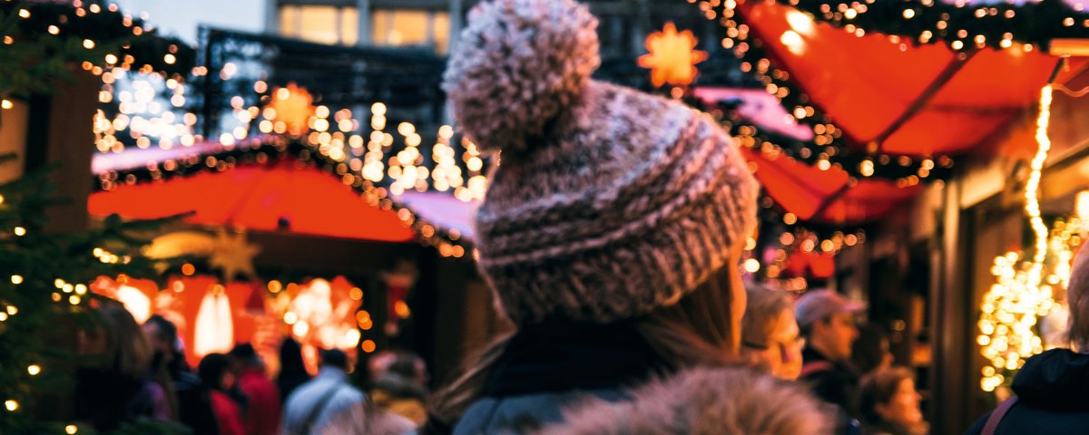 Eine Frau in Winterkleidung mit Mütze läuft über einen Weihnachtsmarkt in Köln, umgeben von Menschen und festlich geschmückten Ständen mit Lichtern.