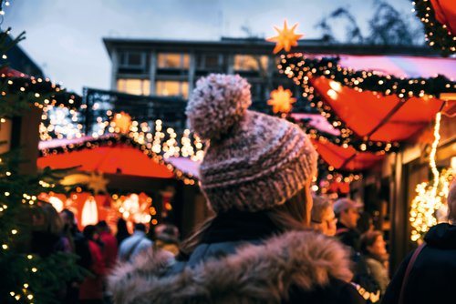 Eine Frau in Winterkleidung mit Mütze läuft über einen Weihnachtsmarkt in Köln, umgeben von Menschen und festlich geschmückten Ständen mit Lichtern.