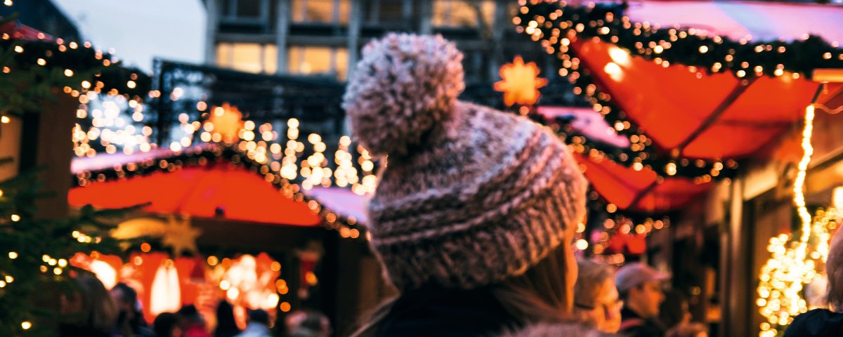 Eine Frau in Winterkleidung mit Mütze läuft über einen Weihnachtsmarkt in Köln, umgeben von Menschen und festlich geschmückten Ständen mit Lichtern.