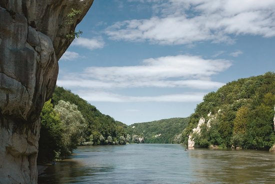 Blick auf die Donau auf der Kataraktenstrecke mit einer Felswand links im Vordergrund und Kanufahrern auf dem Fluss, umgeben von bewachsenen Hügeln unter bewölktem Himmel.