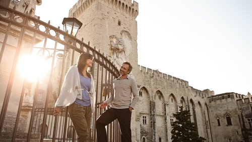 Ein Paar steht in der Altstadt von Avignon vor einem Zaun an einem historischen Gebäude. Im Hintergrund geht die Sonne unter.