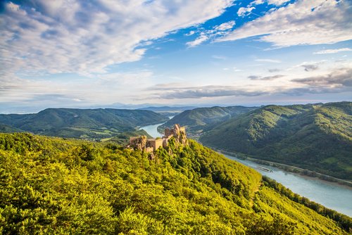 Eine malerische Flusslandschaft in der Wachau mit bewachsenen Hügeln, einem Schloss und einem vorbeifahrenden Flusskreuzfahrtschiff auf der Donau.