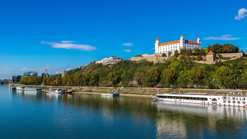 Blick auf die Donau mit mehreren Schiffen und der Burg Bratislava auf einem bewaldeten Hügel unter klarem blauem Himmel