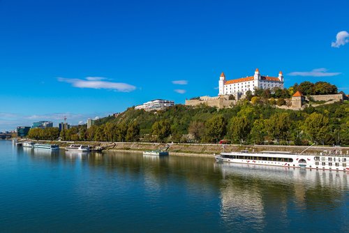 Blick auf die Donau mit mehreren Schiffen und der Burg Bratislava auf einem bewaldeten Hügel unter klarem blauem Himmel