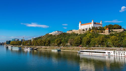 Blick auf die Donau mit mehreren Schiffen und der Burg Bratislava auf einem bewaldeten Hügel unter klarem blauem Himmel