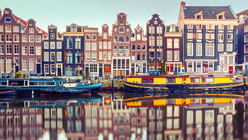 Blick auf Amsterdams Grachten mit historischen Gebäuden, Booten auf dem Wasser und Fahrrädern am Ufer bei Sonnenuntergang.