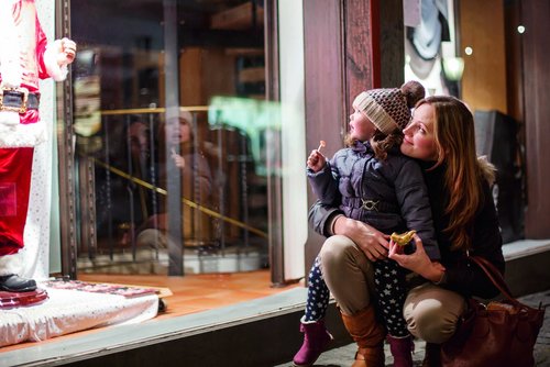 Mutter und Kind schauen gemeinsam ein Schaufenster mit einer Weihnachtsmannfigur an, das Kind hält einen Lutscher und die Mutter ein Stück Gebäck.