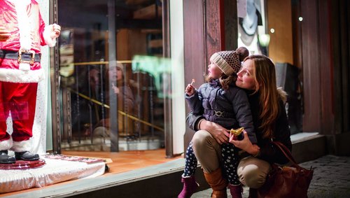 Mutter und Kind schauen gemeinsam ein Schaufenster mit einer Weihnachtsmannfigur an, das Kind hält einen Lutscher und die Mutter ein Stück Gebäck.