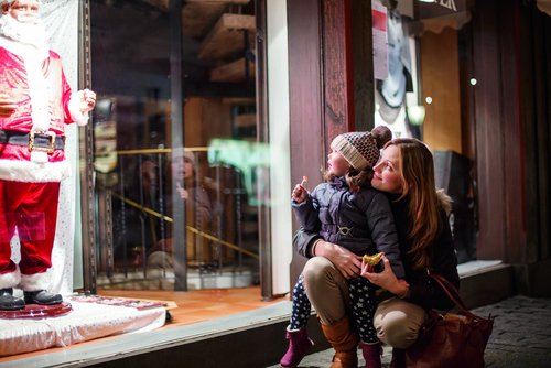 Mutter und Kind schauen gemeinsam ein Schaufenster mit einer Weihnachtsmannfigur an, das Kind hält einen Lutscher und die Mutter ein Stück Gebäck.