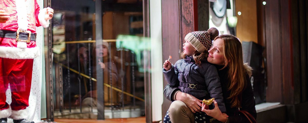 Mutter und Kind schauen gemeinsam ein Schaufenster mit einer Weihnachtsmannfigur an, das Kind hält einen Lutscher und die Mutter ein Stück Gebäck.