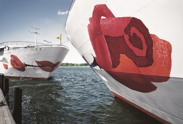 Nahaufnahme eines Flusskreuzfahrtschiffs der A-ROSA Flotte mit markanter Rose auf dem Bug im Vordergrund. Gegenüber steht ein zweites A-ROSA Schiff mit Blick auf den Bug.